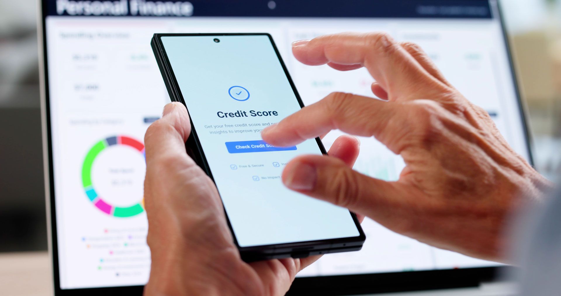 Hands holding a smartphone with a financial app open, a laptop with financial charts in background.