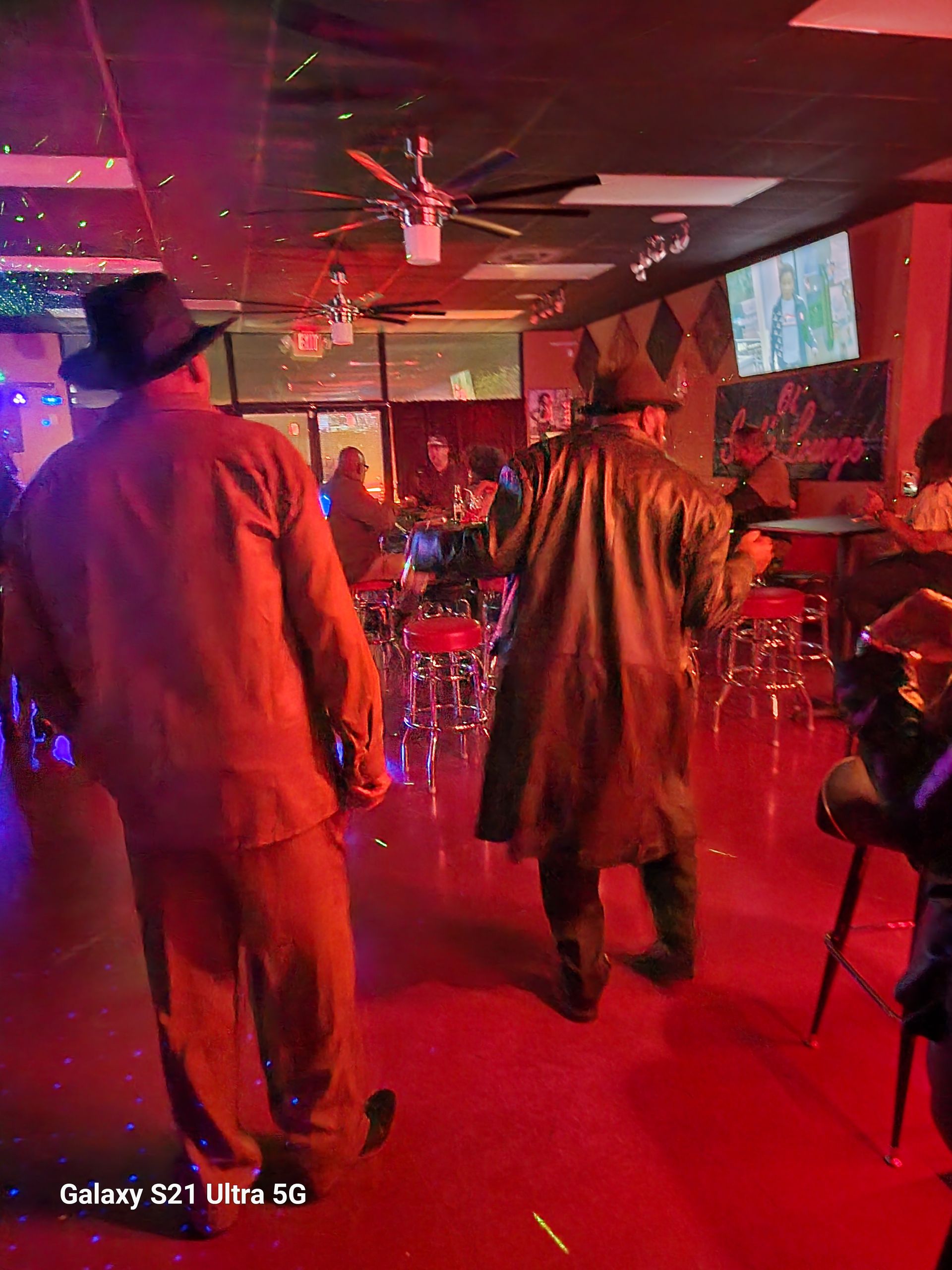 Two men in hats and coats dance in a dimly lit bar with red floors and disco lights.