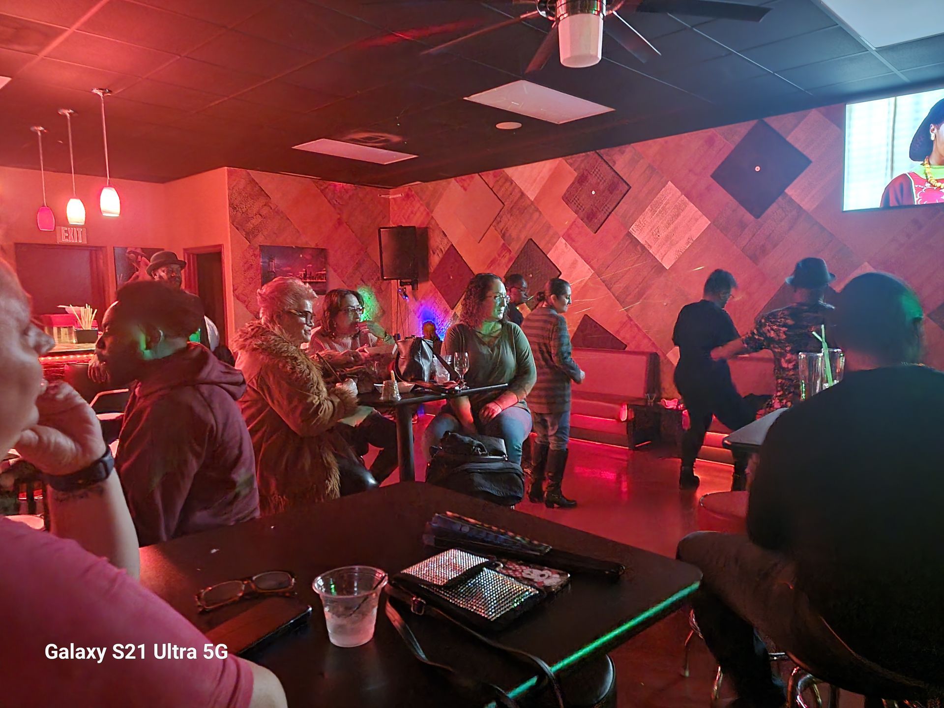 People in a bar with red lighting; some at tables, others at the bar, with a TV screen.