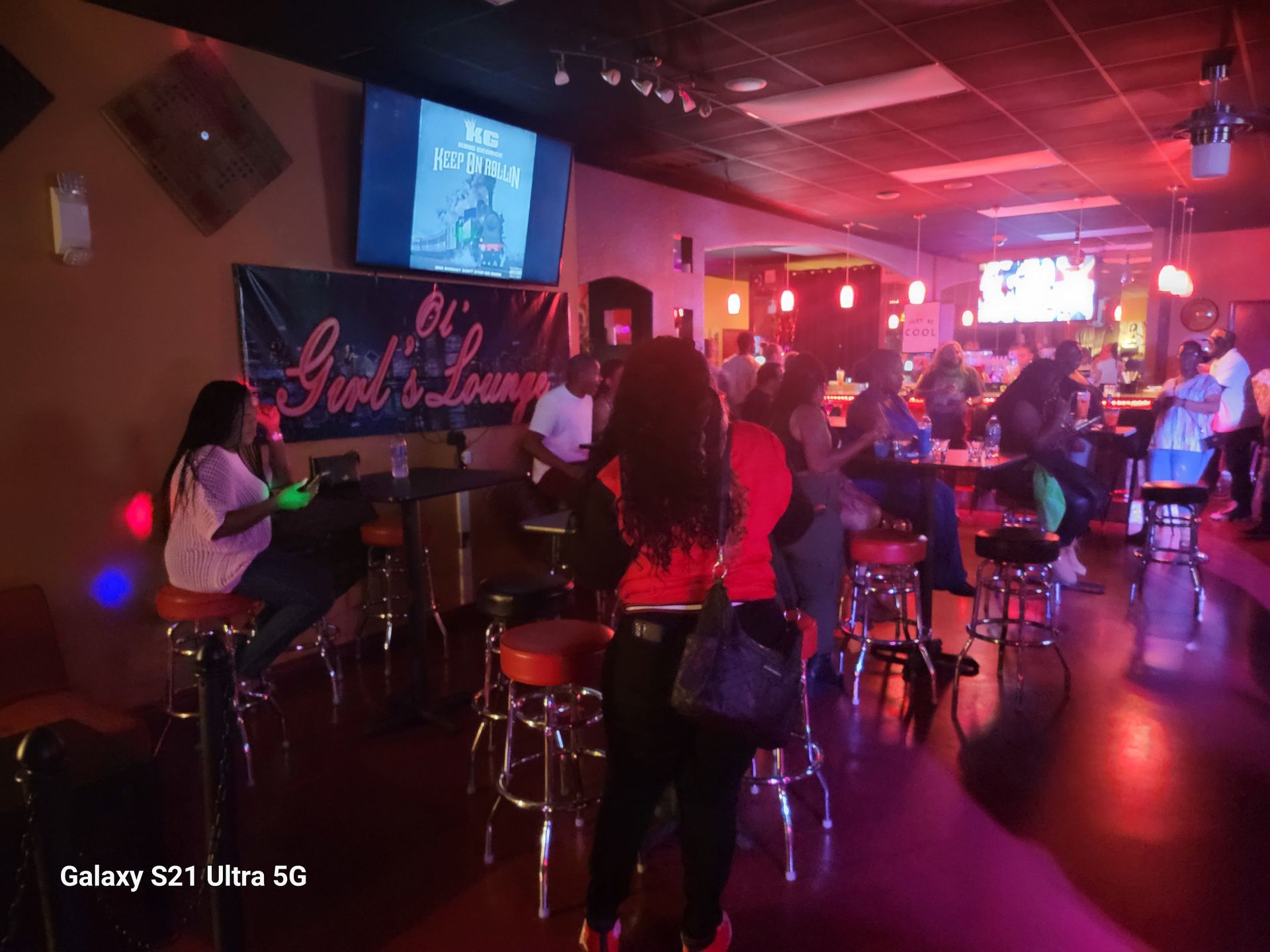 Inside a dimly lit bar, patrons seated at stools, socializing, and watching TVs; red and blue lights.