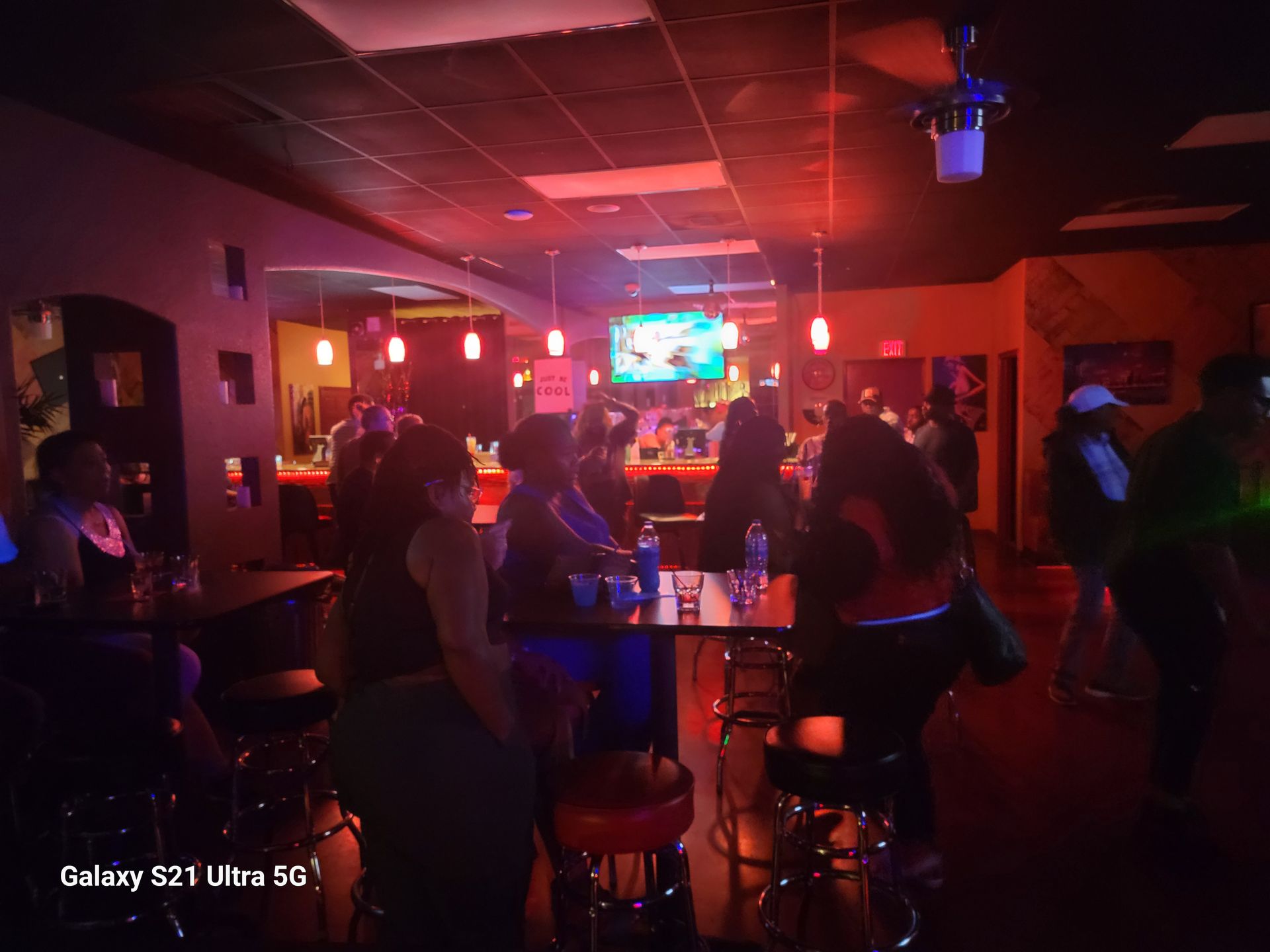 People in a dimly lit bar with red lighting. Some sit at tables, others near the bar, a TV is visible.