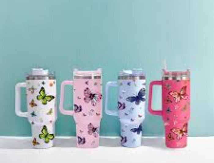 Cuatro vasos térmicos con diseño de mariposas en blanco, rosa, azul y rojo, con asas y sobre un fondo verde azulado.