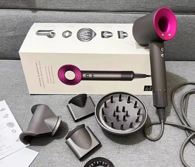 Secador de pelo Dyson con accesorios, caja y manual sobre una superficie gris. El secador es de color gris oscuro y fucsia.