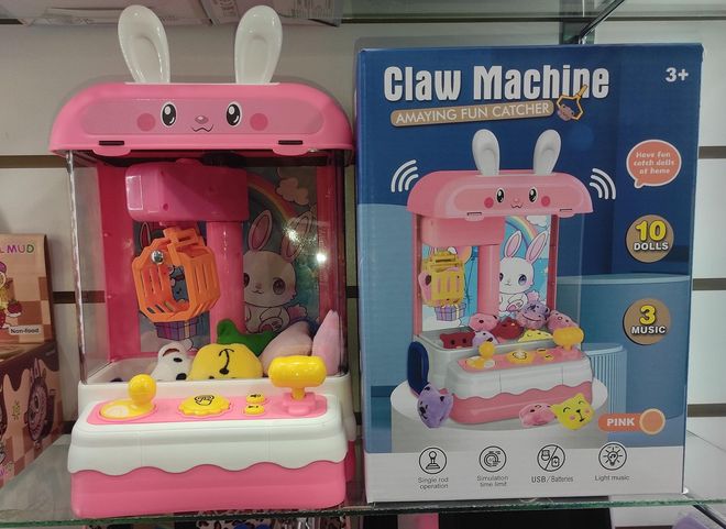 Juguete máquina de garras de conejito rosa junto a su caja, con premios en su interior y expositor colorido.