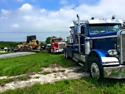 Blue semi truck and red truck on a grassy roadside beside construction equipment under a cloudy sky