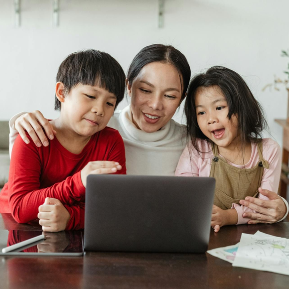 A parent sits at a table with two children, all smiling while looking at a laptop screen.