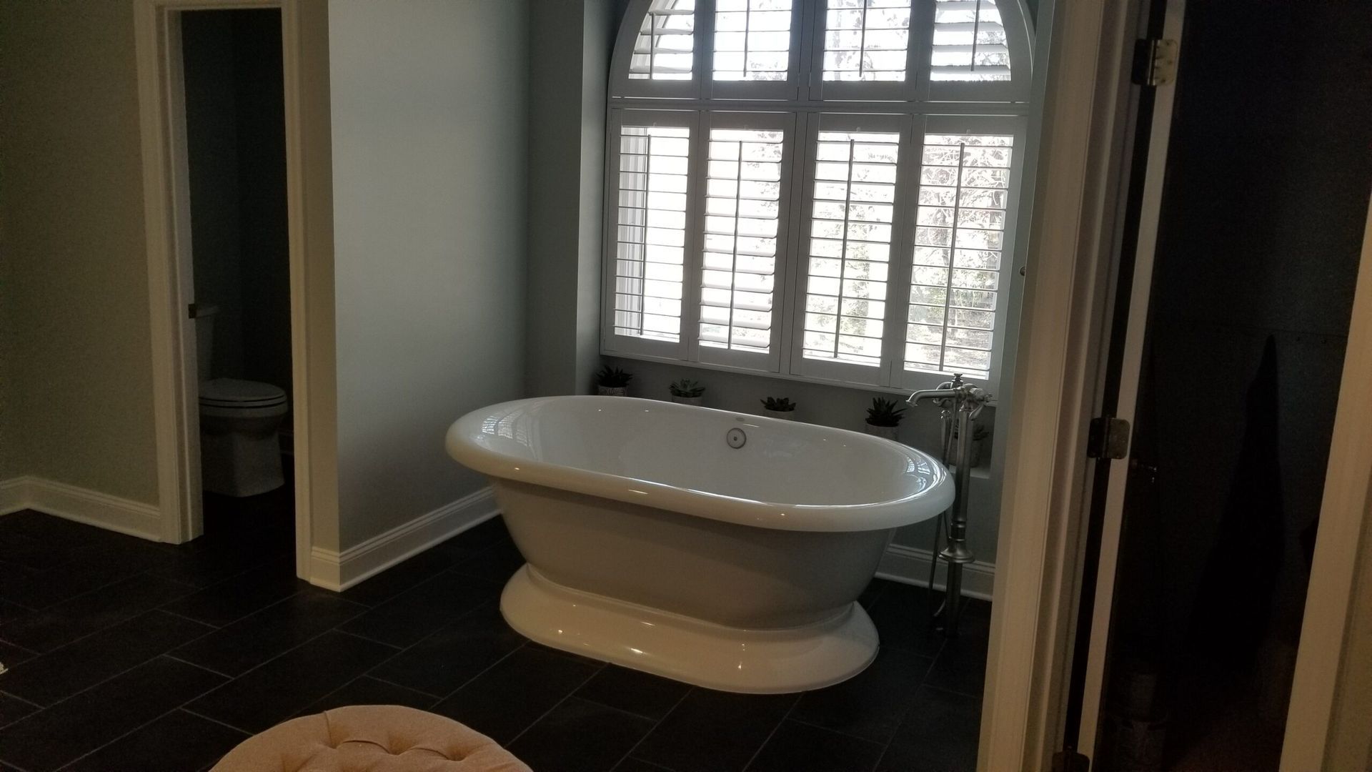 A clawfoot bathtub sits in front of a window with shutters in a bathroom. The bathroom has dark tile floors and light green walls.