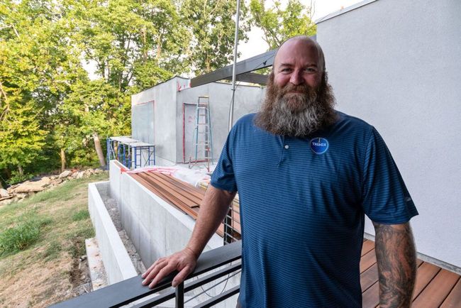 A man with a beard is standing on a balcony in front of a building under construction.