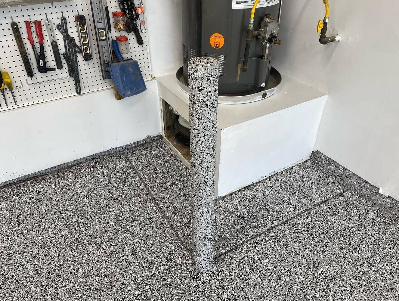 Garage corner with water heater and tools on pegboard, featuring speckled flooring.