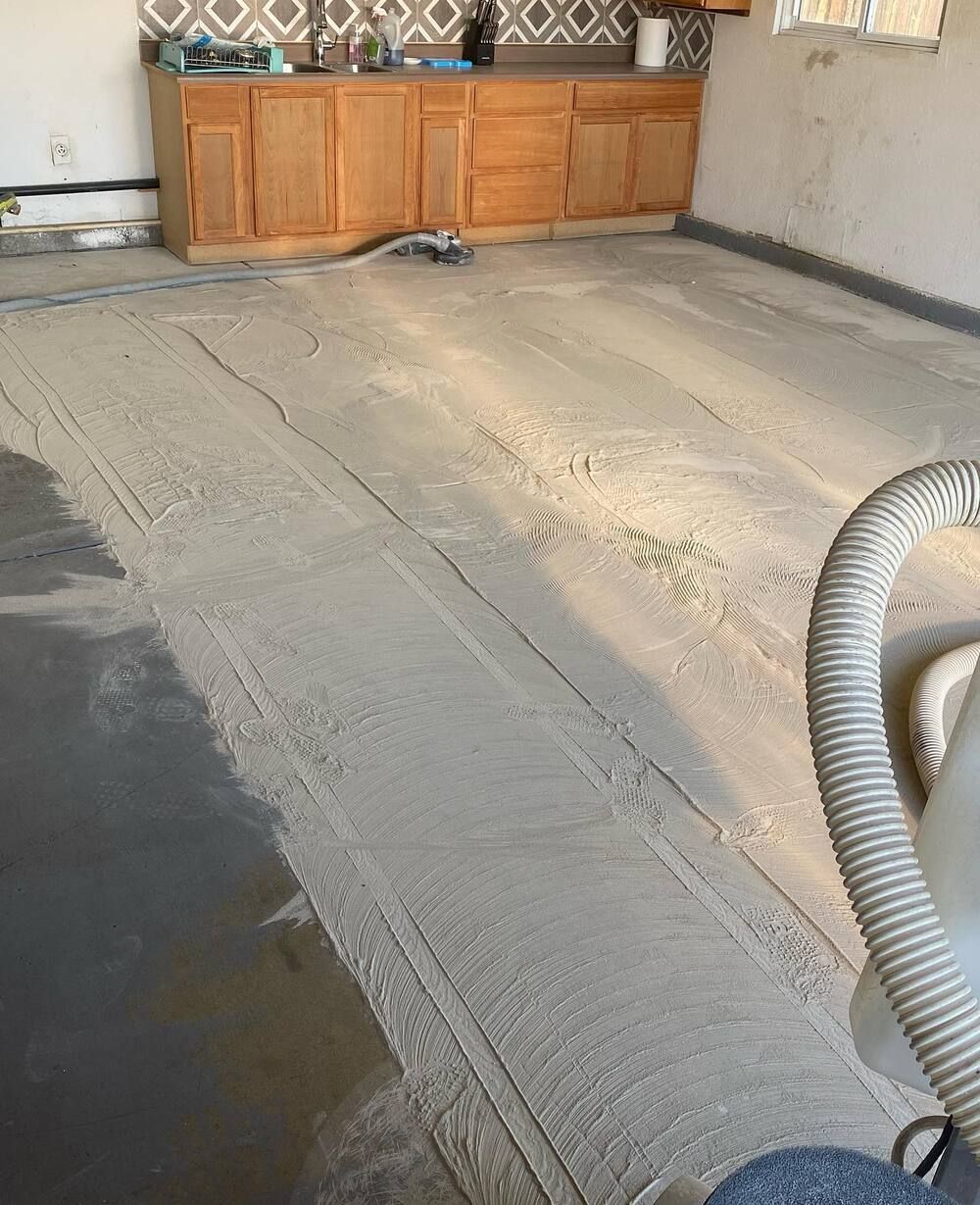 A gray concrete floor being resurfaced in a kitchen setting. A hose and cabinets are visible.