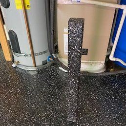 Black, speckled foam roller stands upright on a speckled floor, near water heaters and pipes.