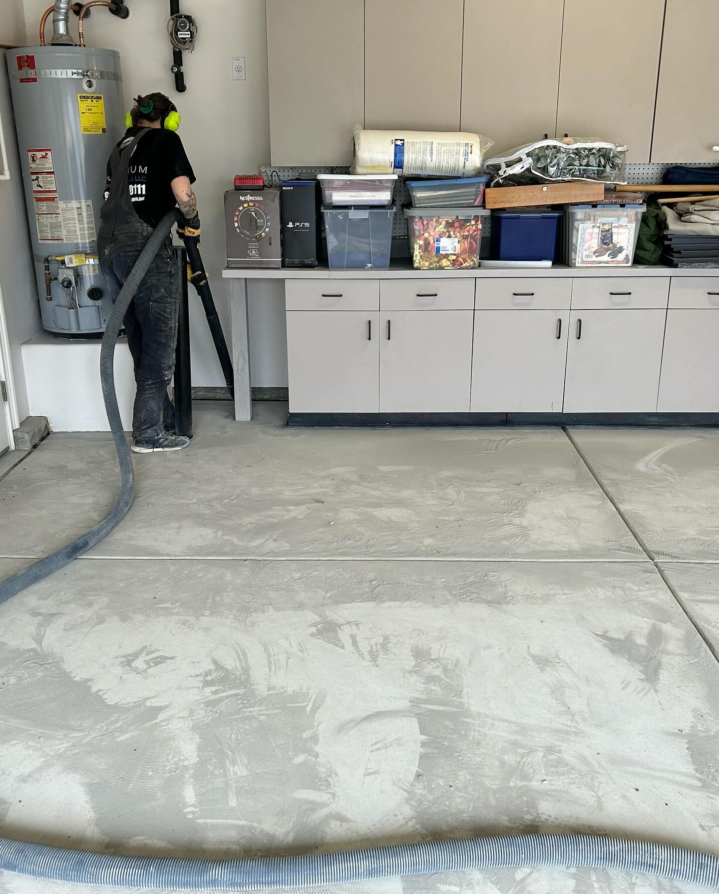 Person using a hose to clean a concrete garage floor near a water heater and storage cabinets.