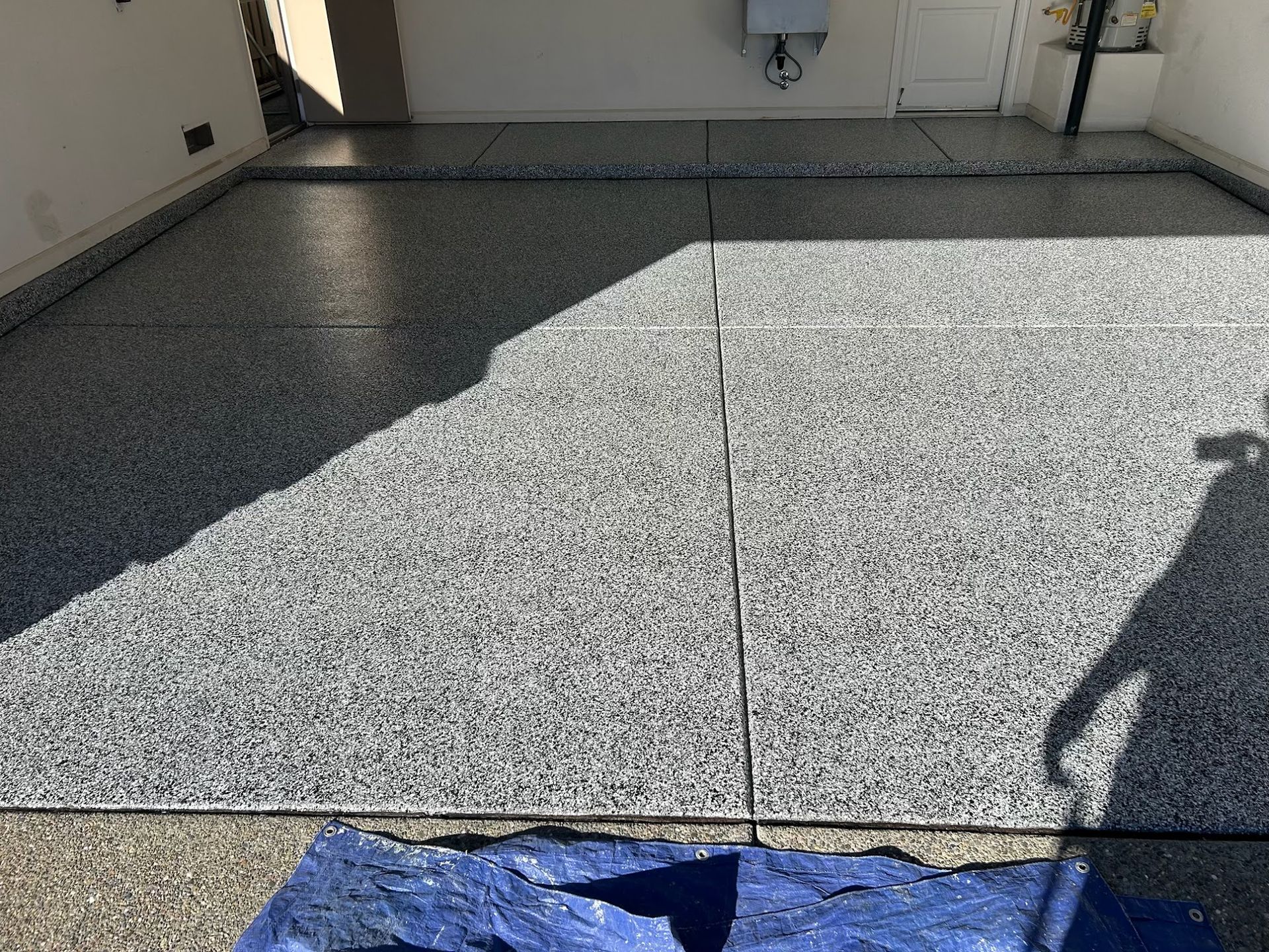 Epoxy-coated garage floor with gray and black flecks. Blue tarp in front, sunlight across the space.