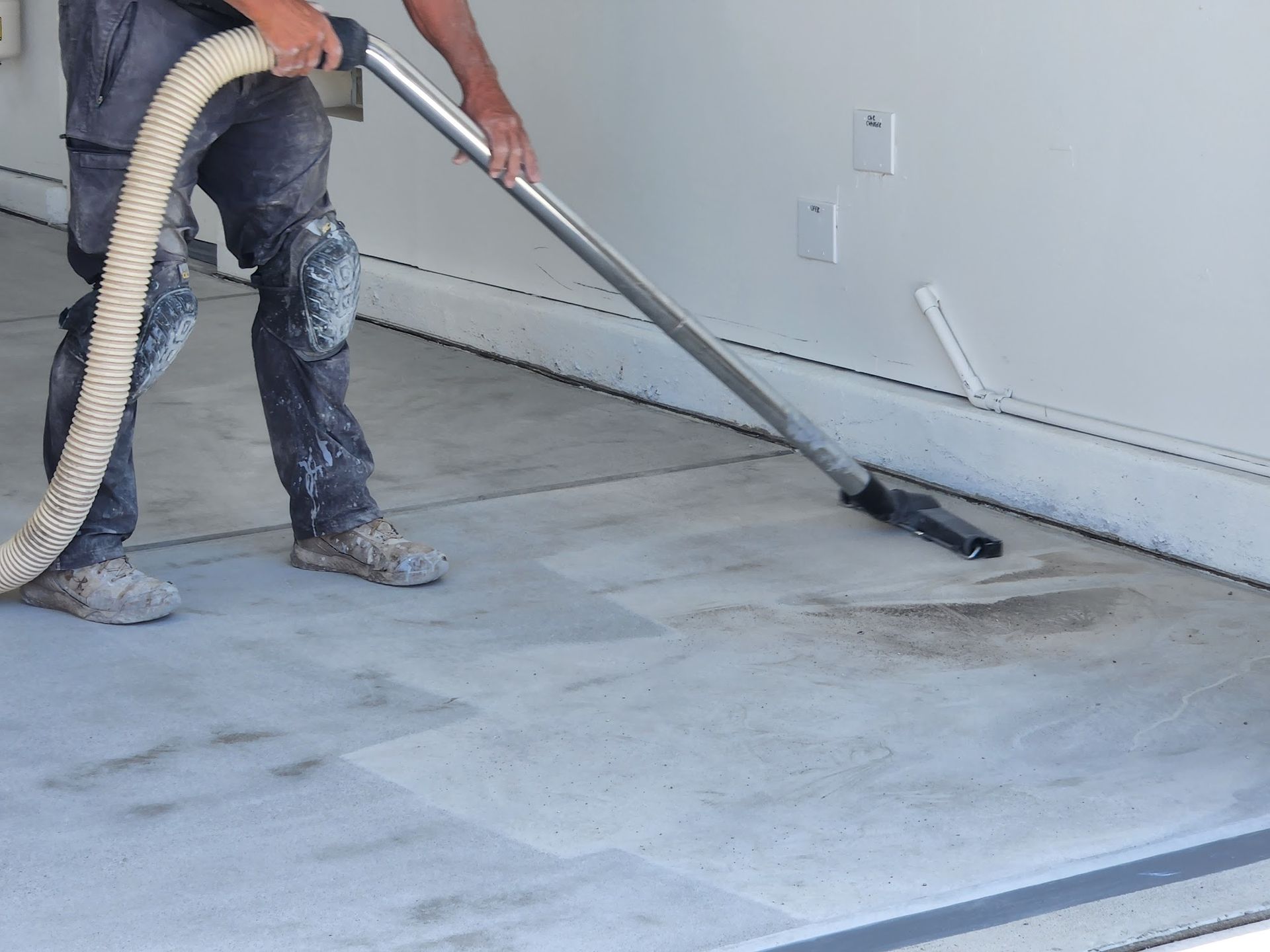 Person vacuuming a concrete floor in a garage; hose, metal wand, and debris visible.