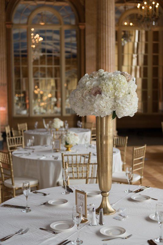 Elegant wedding reception table with gold vase floral centerpiece. Round tables, gold chairs, mirrored windows.