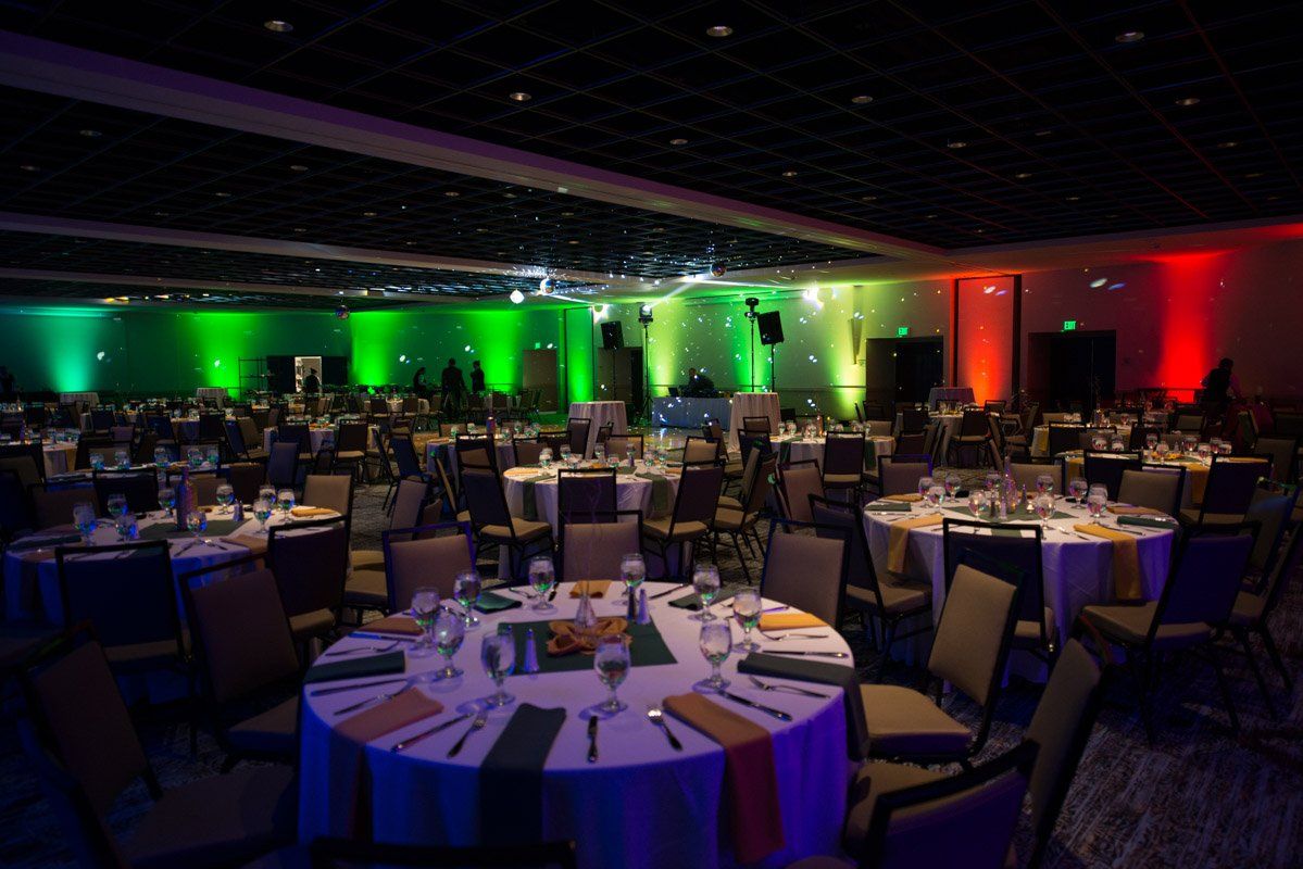 A banquet hall with round tables set for a formal event, lit by green and red stage lights.