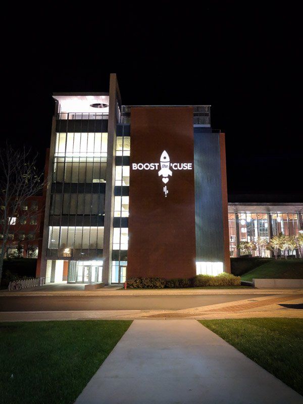 Night view of the Boost House building with the rocket logo illuminated. Brick facade, glass windows, and a grassy lawn.