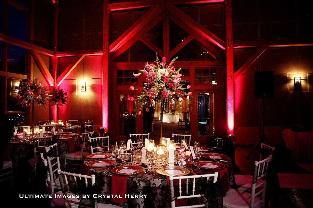Red-lit wedding reception with round tables, floral centerpieces, and white chairs in a rustic building.