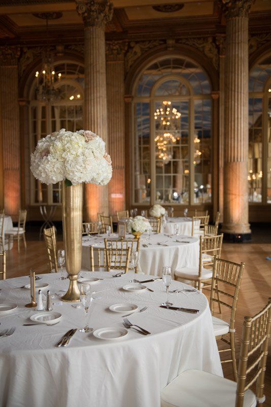 Elegant wedding reception tables, golden chairs, tall floral centerpieces, in a grand ballroom.