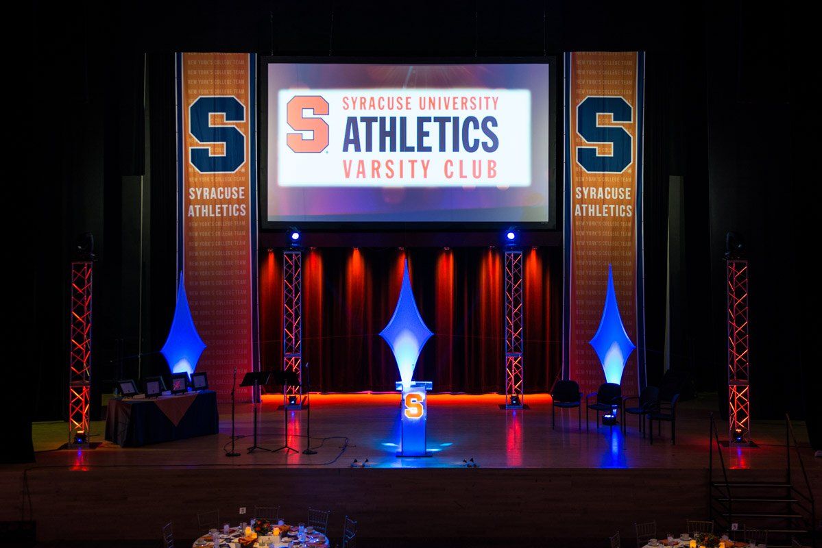 Stage set with Syracuse University Athletics Varsity Club logo and orange/blue lighting.