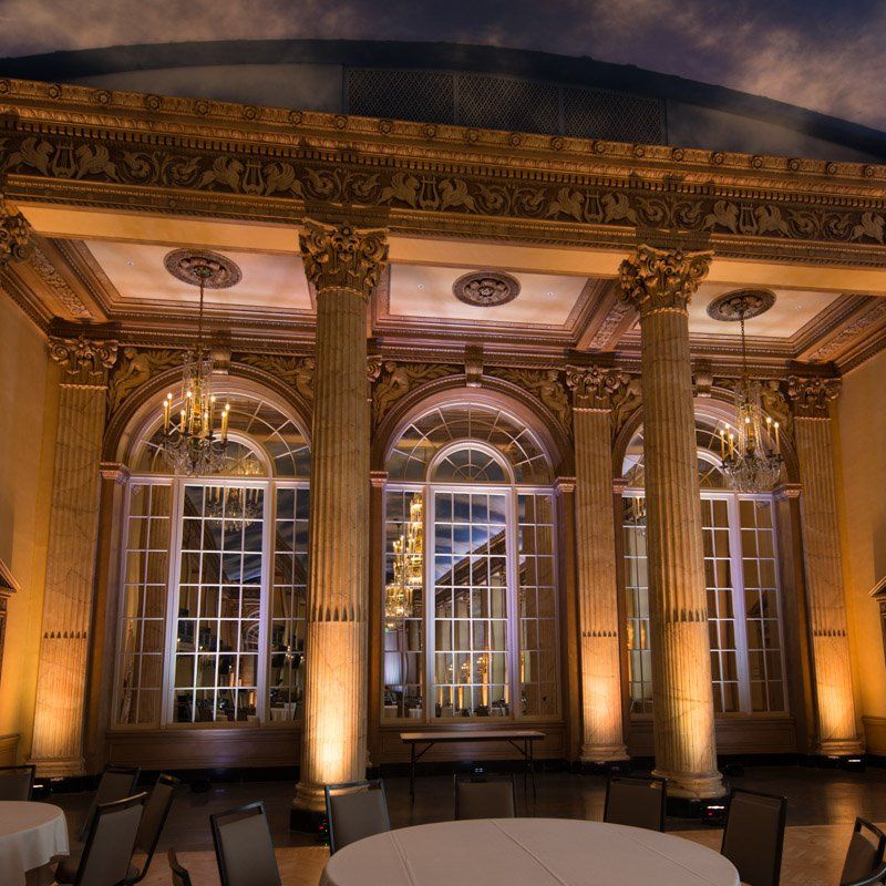 Elegant ballroom with tall columns, arched windows, chandeliers, and round tables.