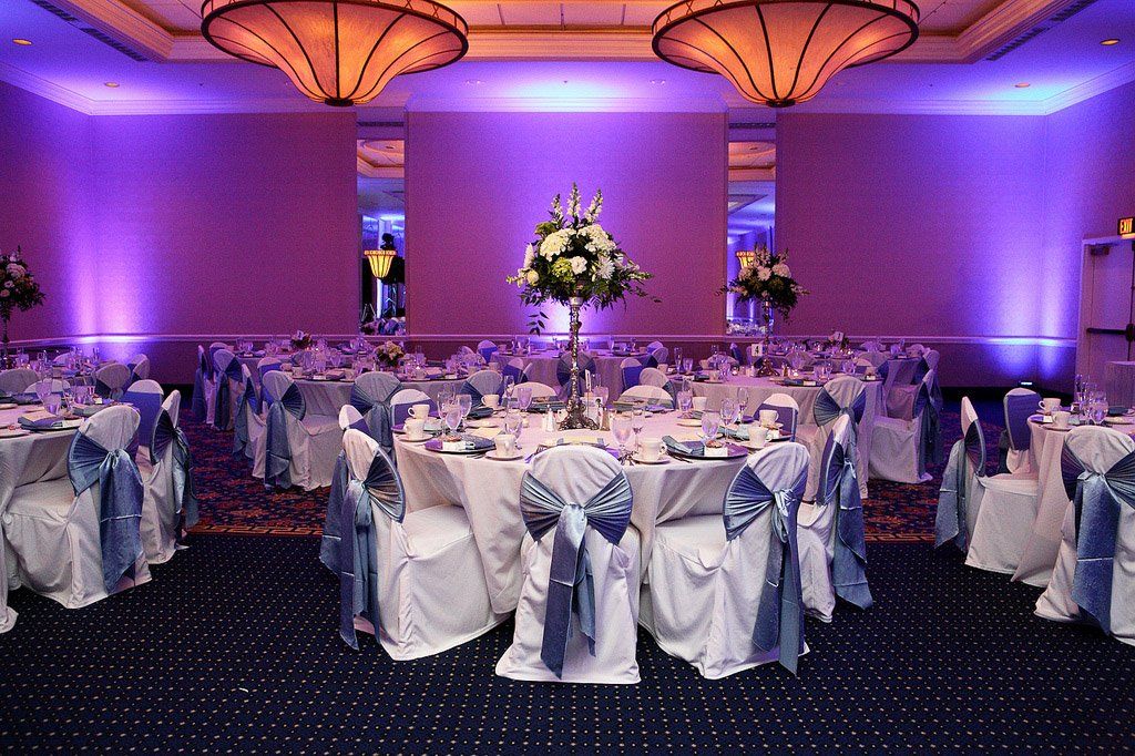 Elegant banquet hall with round tables set for a formal event; white chairs, floral centerpieces, and blue ambient lighting.