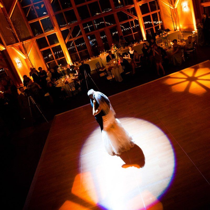 Couple dances on a wooden dance floor in a spotlight at a wedding reception. Warm lighting illuminates the venue.