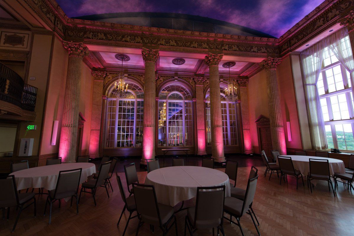 Ornate ballroom with pink lighting, round tables, and large windows.