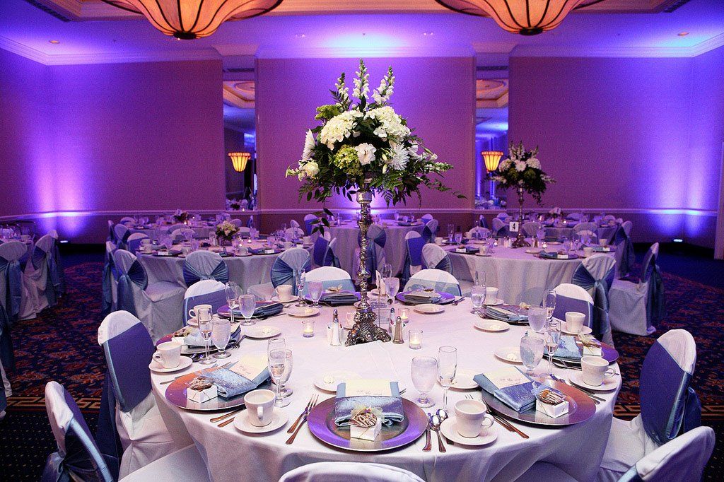 Formal dining room set for an event; round tables with white and purple decor; flowers, and blue up lighting.
