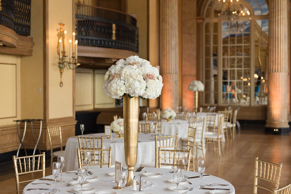 Elegant ballroom with gold accents, round tables with white linens, floral centerpieces.