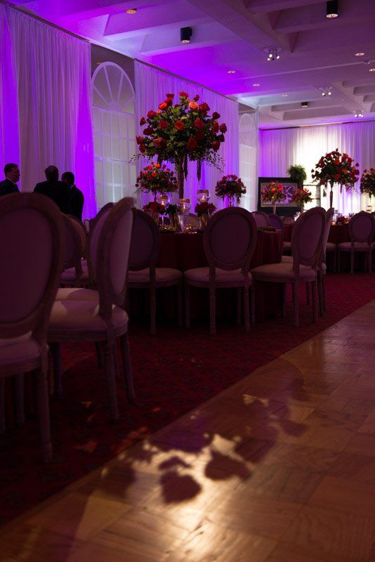 Elegant banquet hall with round tables, floral centerpieces, purple lighting, and white chairs.