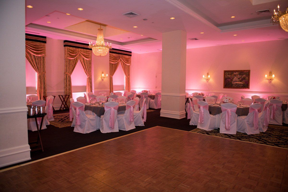 A banquet hall with round tables set for a reception. Pink lighting, white chairs, and dance floor.