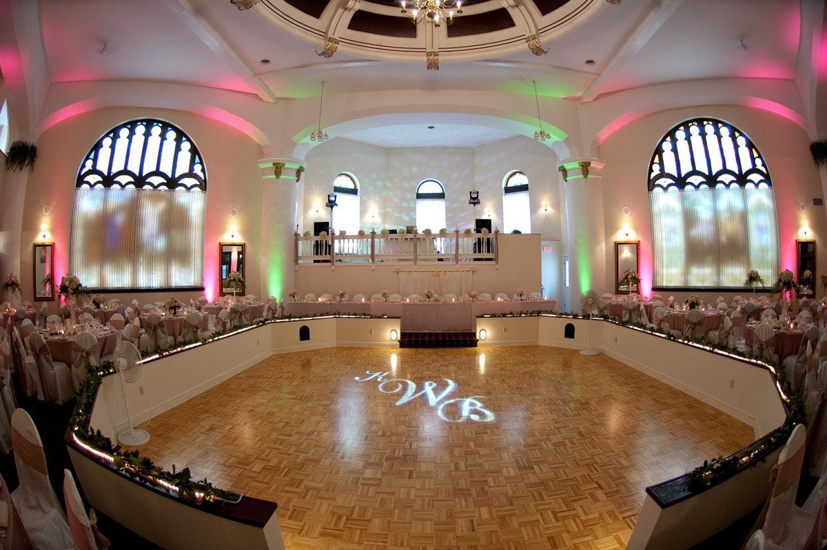 A large, ornate ballroom with a wooden dance floor and stage, featuring tables, chairs, and colorful lighting.