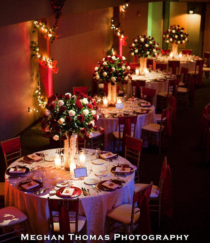 Elegant reception hall with round tables set for dinner; red and white floral centerpieces, soft lighting.