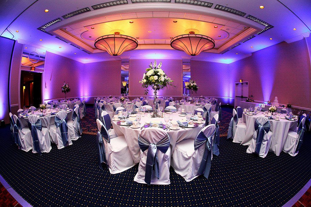 Reception hall with round tables set for an event. Tables have white linens with blue sashes, bathed in purple light.