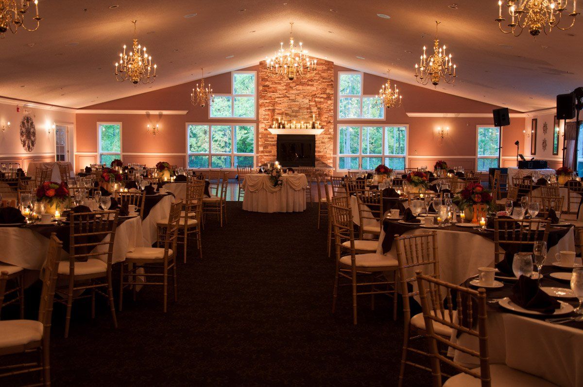 Reception hall with tables set for a formal event; gold chairs, centerpieces, chandeliers, and a brick fireplace.