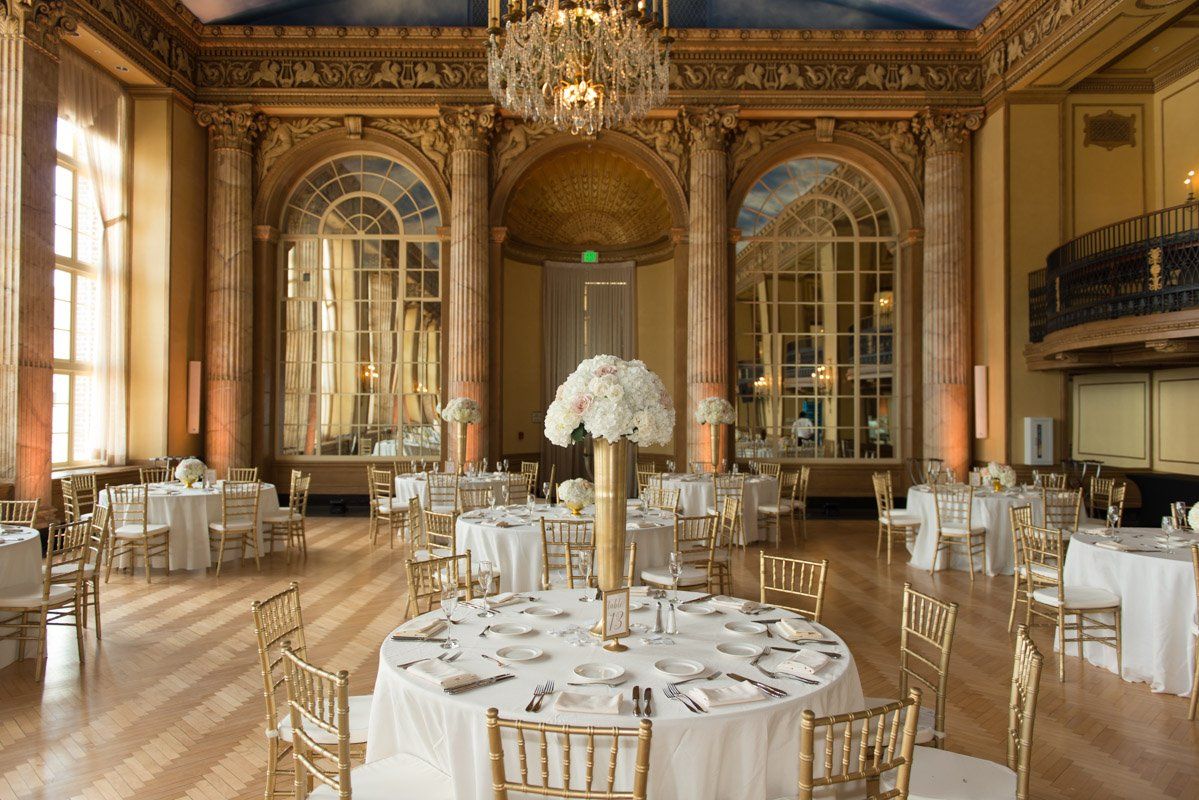 Elegant ballroom with round tables, gold chairs, and large mirrors. A floral centerpiece adorns one table.