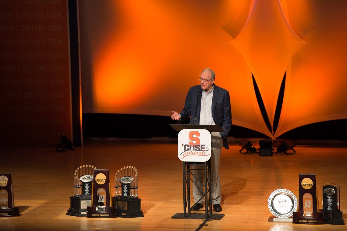 Man speaking at podium with Syracuse trophies. Orange stage backdrop.