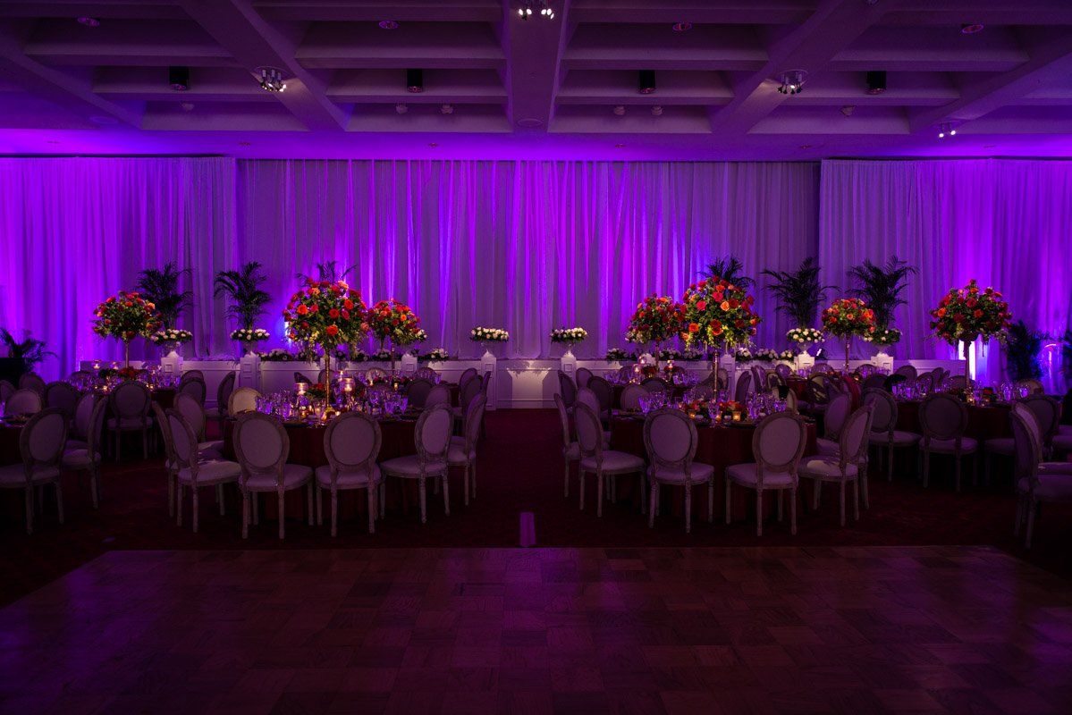 A wedding reception hall with purple lighting, round tables with floral centerpieces, and formal chairs.