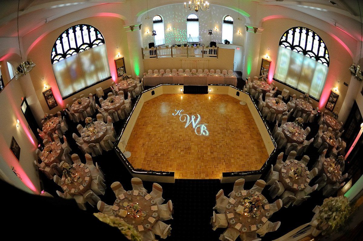 Reception hall with round tables set for guests. A large dance floor features initials, illuminated arches.