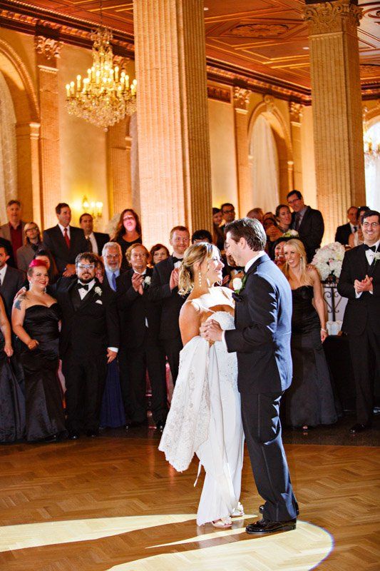 Bride and groom dancing at their wedding reception, guests watching; ornate ballroom setting.