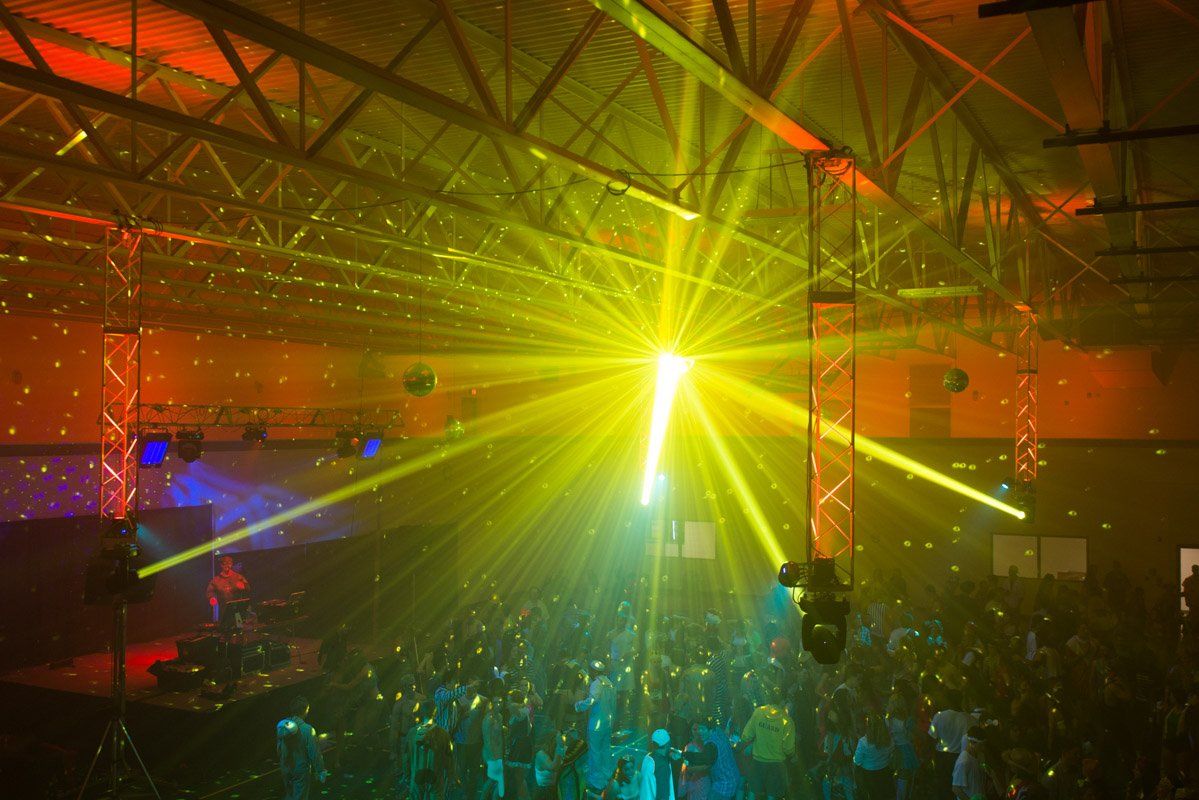 People at a concert dance under yellow and green laser lights inside a large building.
