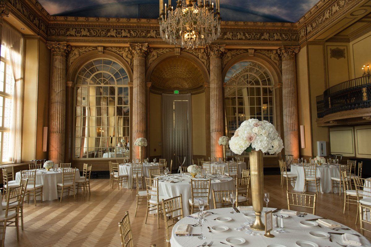 Elegant ballroom set for a wedding reception with round tables, gold chairs, and large windows.