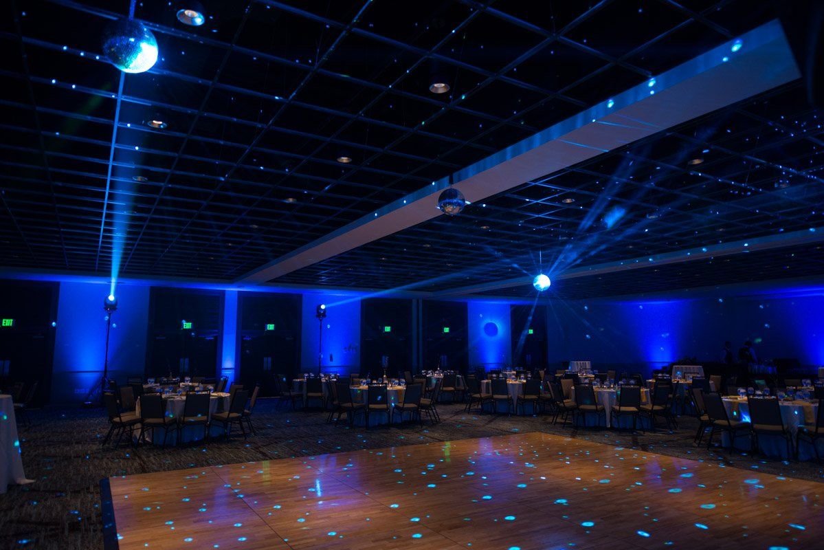 A ballroom with blue lighting and a mirrored disco ball. Tables are set, and the dance floor reflects blue lights.
