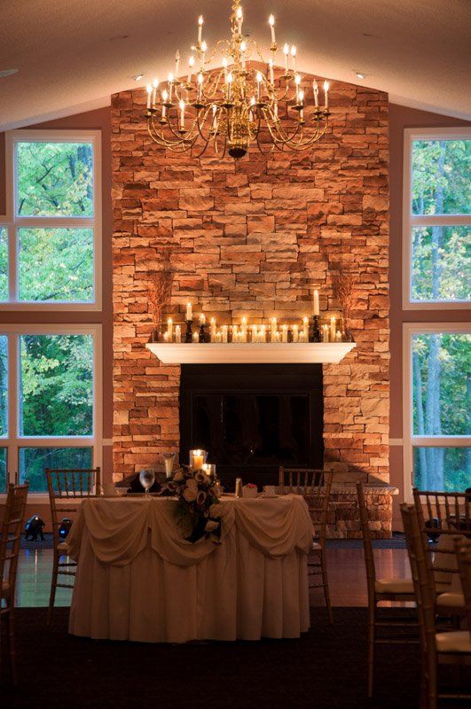 Wedding reception room with a stone fireplace, candelabra, and windows overlooking trees. Tables set for guests.