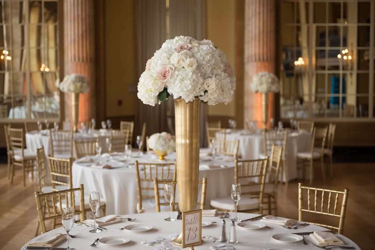 Wedding reception with round tables, gold chairs, floral centerpieces, and ornate columns.