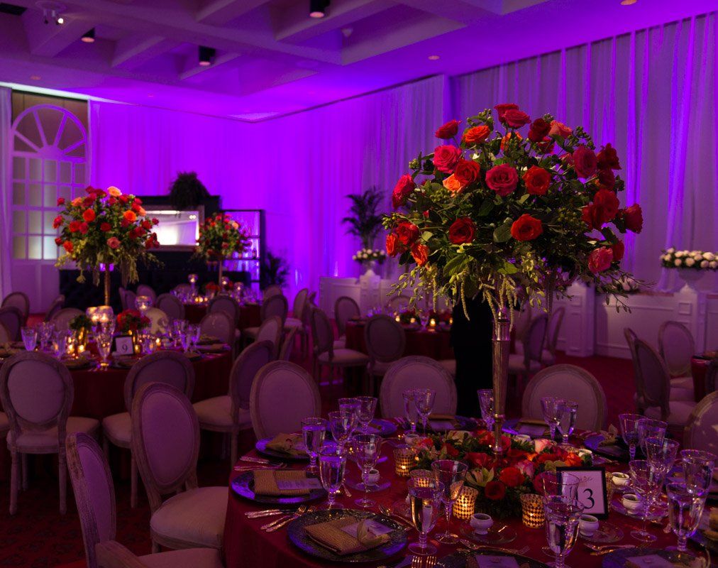 Elegant banquet hall with tables, chairs, and tall floral centerpieces; bathed in purple light.