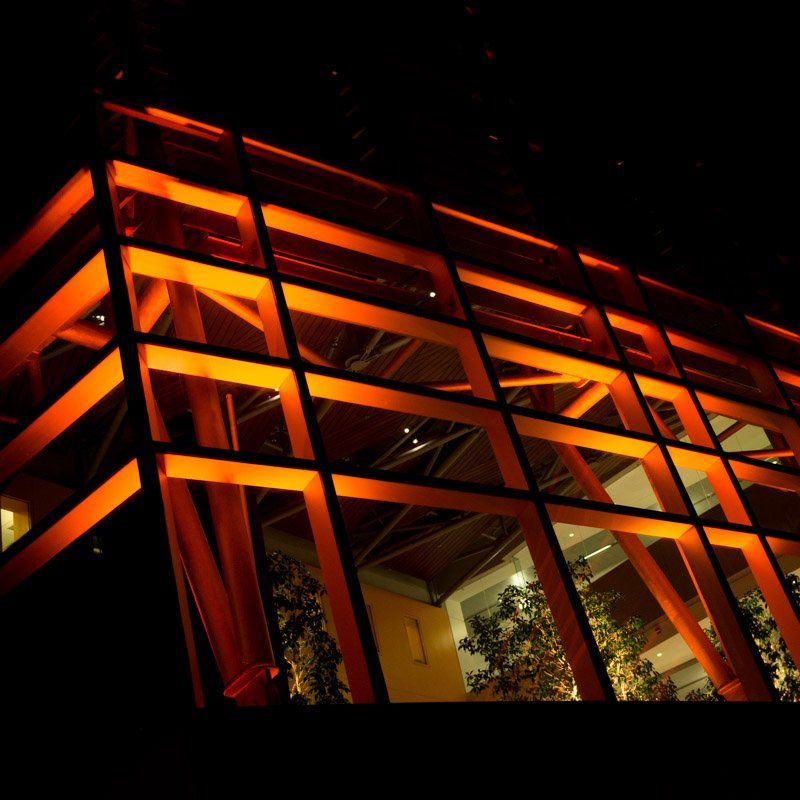 Orange-lit modern building at night. Large windows, visible wooden beams, dark background.