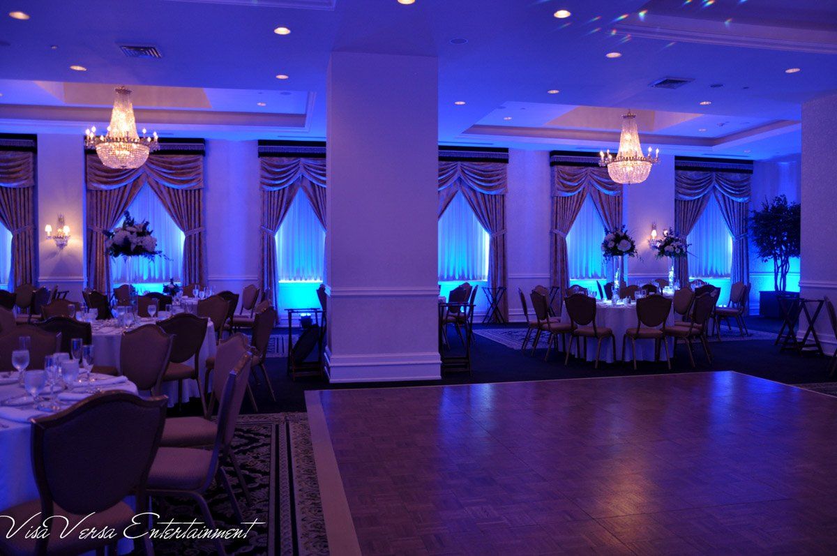 Formal ballroom with blue up-lighting, chandeliers, round tables, and a dance floor.