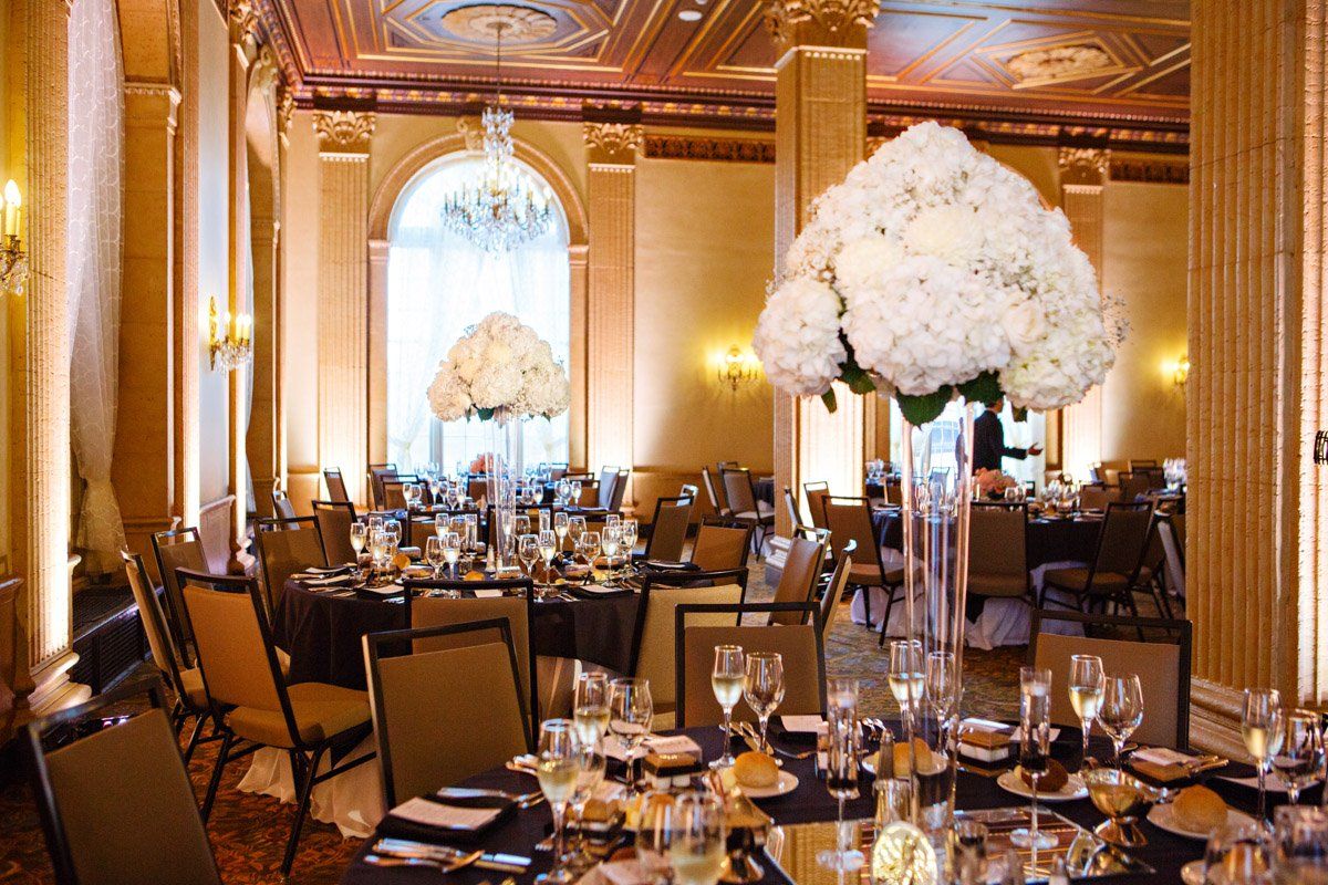 Elegant ballroom with tables set for a formal event. White floral centerpieces, dark linens.